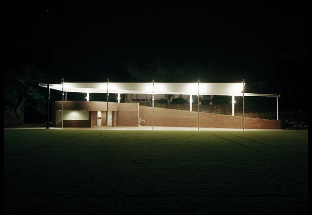 A lightweight steel roof is tethered above the brick pavilion, its underside emphasized by uplighting.