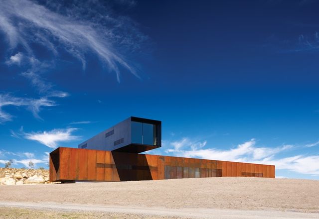 A Corten block is set athwart the hill, with the black upper floor sitting at a right angle across it.