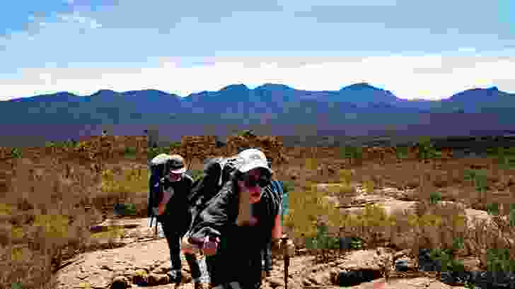 Hikers traverse mountainside rocky slabs, guided by yellow markers that are fixed to rocks at intervals.